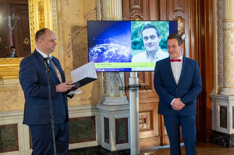 Zwei Männer in Anzügen stehen vor dem TV-Bildschirm in einem goldenen Raum