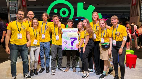 A group photo. 11 young people pose together. All are waring the same yellow t-shirts. The person in the middle holds a poster that says "WunderBand"