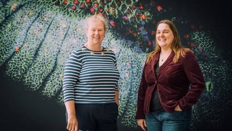 two people standing next to each other smiling. Behind them, there is a colorful background of a painting.