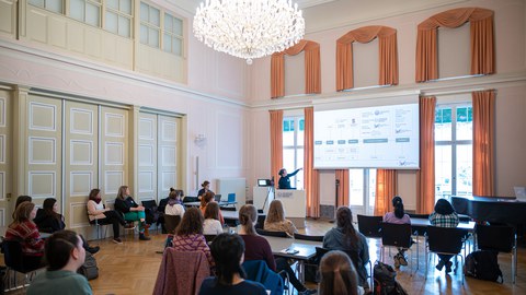 Women sitting in an old-school ballroom room. A person stands in front and gives a talk with the slides projected on the beamer. 