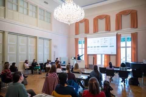 Frauen, die in einem Ballsaal der alten Schule sitzen. Eine Person steht vorne und hält einen Vortrag, wobei die Folien auf den Beamer projiziert werden. 