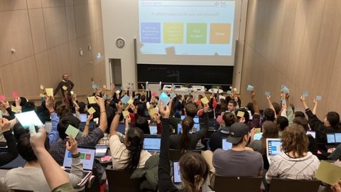 Studierende in einem Hörsaal sehen eine Präsentation mit farbigen Rechtecken, die Text enthalten. Die Studierenden halten in einer Hand Haftnotizen in verschiedenen Farben, die den Rechtecken entsprechen, hoch (blau, gelb, grün, orange) 