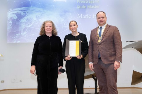 Zwei Frauen und ein Mann lächeln und posieren für ein Foto. Die Frau in der Mitte hält eine Mappe mit einer Urkunde. Der Mann auf der rechten Seite trägt eine goldene Amtskette.nten. 