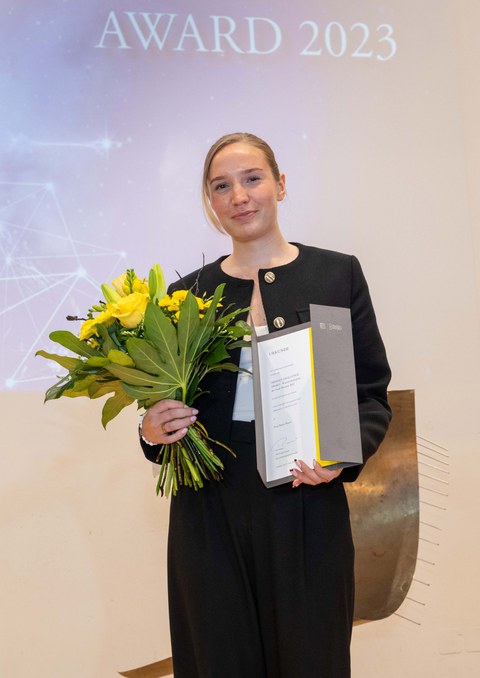 Eine Frau posiert für ein Foto, in der Hand einen Strauß mit gelben Blumen und eine Urkunde. Im Hintergrund ist der Text "AWARD 2023" an die Wand projiziert.