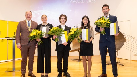 Three men and two women posing for a photo. The man on the left has a golden chain of office. The remaining four people hold buquets with yellow flowers and certificates. The text "DRESDEN EXCELLENCE AWARD 2023" is projected on the wall behind.