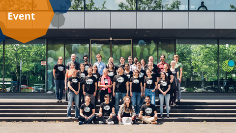 Ein Gruppenfoto. Etwa 30 Personen stehen in vier Reihen auf einer Treppe vor einem Gebäude. Die meisten Personen tragen passende T-Shirts. Das Gebäude hat Glaswände, die die grünen Bäume und den blauen Himmel widerspiegeln.