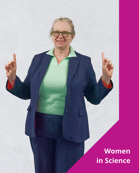Ein Foto mit grafischen Elementen. Eine lächelnde Person steht mit den Händen auf Schulterhöhe und den Zeigefingern nach oben gerichtet. Auf der rechten Seite befindet sich ein rosa visuelles Element mit dem Text „Women in Science” (Frauen in der Wissenschaft) am unteren Rand.