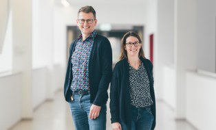 A Groupphoto. Two people smiling standing in a hallway.