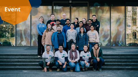 Ein Foto mit grafischen Elementen. Ein Gruppenfoto von 20 Personen, die in vier Reihen auf einer Treppe vor einem Gebäude stehen. Die Personen blicken in die Kamera und lächeln. Die Personen in der ersten Reihe tragen sichtbare Namensschilder, die an ihren identischen grauen T-Shirts befestigt sind. Im Hintergrund ist eine bunte Glastür zu sehen. In der oberen linken Ecke befinden sich bunte Achtecke mit dem Wort „Event”. 