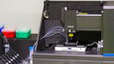 A photo. Laboratory equipment sits on a metallic surface. The dark gray and black equipment has an open top showing wires. To the left, a plastic case holds clear tubes.