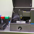 A photo. Laboratory equipment sits on a metallic surface. The dark gray and black equipment has an open top showing wires. To the left, a plastic case holds clear tubes.