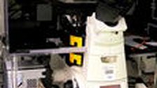 A photo. The image shows a white microscope with black ocular lenses on a black table. On its bottom there are black knobs, a digital display with red numbers, and two yellow labels.