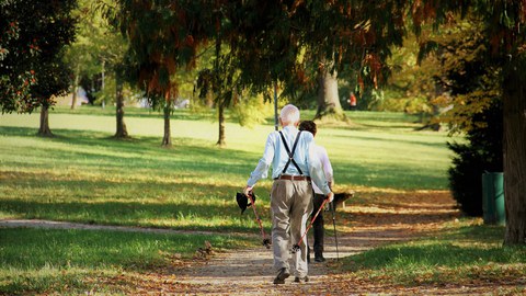 Ein Foto von einem Weg in einem begrünten Park mit vielen Bäumen. Auf dem Weg laufen zwei ältere Menschen hintereinander mit Nordic Walking Stöcken. 