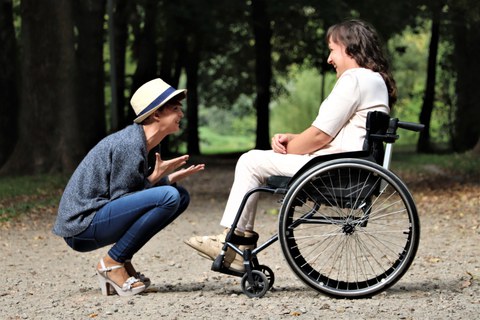 Ein Foto, das zwei Frauen zeigt: die rechte Frau trägt ein helles Oberteil mit farblich passender Hose und Sandalen. Sie hat langes dunkles Haar und sitzt in einem schwarzen Rollstuhl. Vor ihr hockt eine jüngere Frau in einem dunklen Outfit und hellen Sandalen. Sie trägt einen hellen Hut und gestikuliert mit den Händen. Beide Frauen lachen sich an. Im Hintergrund sind verschwommen einige Bäume zu erkennen. 