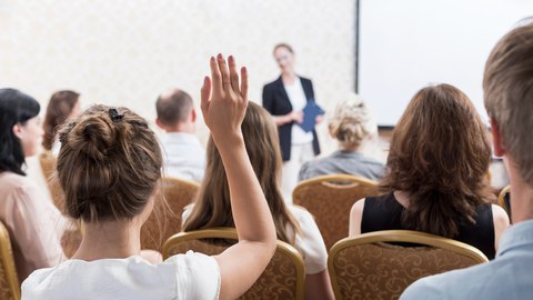 Das Foto zeigt eine Gruppe von Menschen, die sich die Präsentation einer Person anhört. Eine Frau im Vordergrund des Bildes hebt ihren Arm für eine Wortmeldung.