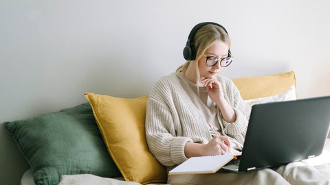 Blonde, junge Frau sitzt auf einem Bett mit einem Laptop auf dem Schoß.