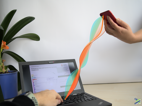 Ein Laptop und ein Mobiltelefon nebeneinander, beide werden jeweils von Händen bedient. Das Handy zeigt auf den Laptop, dazwischen wurde mit grafischen Elementen nachträglich eine Art Welle in div. Farben eingefügt.