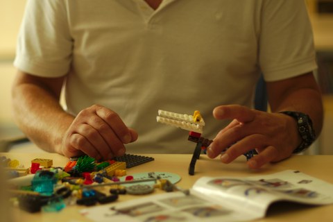 Eine Person sitzt an einem Tisch und baut Lego. Zu sehen ist nur der Oberkörper unterhalb des Kopfes. 