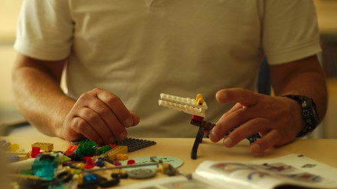 Eine Person sitzt an einem Tisch und baut Lego. Zu sehen ist nur der Oberkörper unterhalb des Kopfes. 