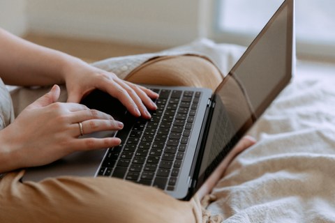 Eine junge Person sitzt auf einem hell bezogenen Bett mit einem Laptop auf dem Schoß