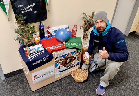 Foto: Mann in Sportkleidung  mit Schokoladenweihnachtsmann in der Hand hockt neben einem kleinen Tisch mit Handtüchern, Schokoladenweihnachstmännern und Tannenzweigen