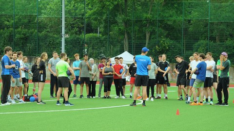 Foto: vor einer Gruppe Personen auf dem Rasen eines Sportplatzes steht ein Mann mit einem blauen Shirt mit dem Rücken zum Betrachter. Auf dem Shirt steht auf dem Rücken "Coach"