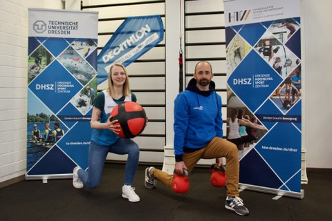 Foto: eine Frau mit einem Medizinball und ein Mann mit roten Kettlebells posieren zwischen Banners des Dresdner Hochschulsportzentrums