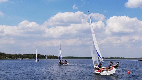 Foto mehrere Segelboote fahren hintereinander über einen See, Himmel mit weißen Wolken im Hintergrund