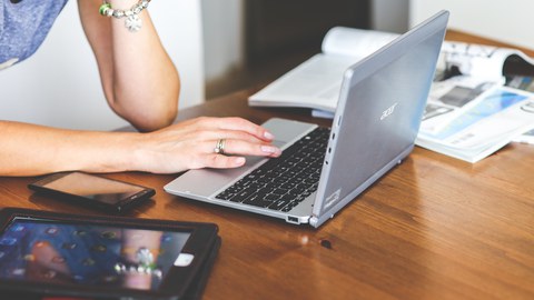 Foto: Schreibtisch mit aufgeklapptem Laptop und daran arbeitendem Menschen