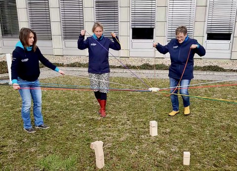 Foto: drei Frauen halten einen Holzklotz mit bunten Fäden in der Luft