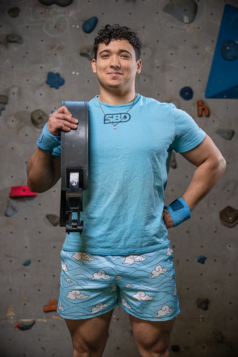 junger Mann in Sportkleidung mit einem Gewichthebergürtel in der Hand steht vor Kletterwand