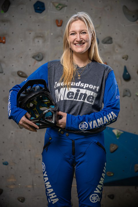 junge Frau in Sportkleidung mit einem Mountainbikehelm unterm Arm steht vor Kletterwand