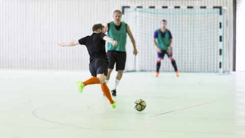 Foto zwei Männer spielen Fußball in der Halle, im Hintergrund sieht man das Tor mit Torwart