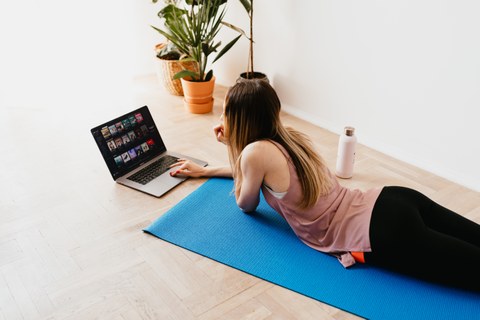 Foto: Junge Frau in Sportkleidung liegt auf einer Yogamatte vor einem Laptop