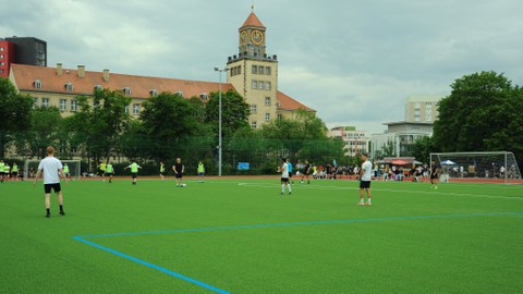 Foto: auf einem Sportplatz mit grünem Rasen spielen Sportler, im Hintergrund ist ein Gebäude mit Uhrenturm zu sehen (Weberbau)