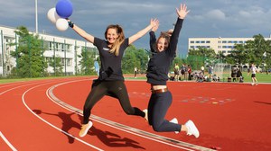 Foto: 2 junge Frauen springen in die Luft, im Hintergrund ist ein Sportplatz mit rotem Boden und weitere Personen zu sehen