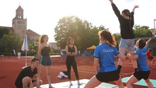 Gruppe von Menschen macht Akrobatik, im Kern der Grupe steht eine Person auf den Oberschenkeln zweier Personen