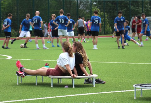 Zwei Frauen machen Pause, auf einem Mini-Trampolin sitzend, im Hintergrund sind Fußballer zu sehen