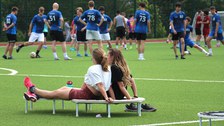 Zwei Frauen machen Pause, auf einem Mini-Trampolin sitzend, im Hintergrund sind Fußballer zu sehen