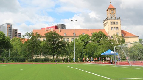 Foto im Vordergrund grüner Sportplatz, auf der Laufbahn dahinter laufende Sportler:innen, dahinter ein großes Gebäude mit Uhrenturm (Weberbau)