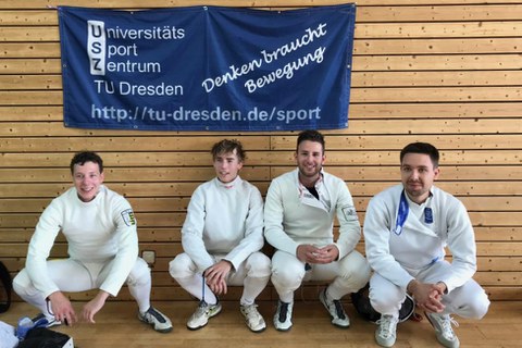 Foto vier Sportler in Fechtkleidung hocken unterhalb eines blauen Banners mit dem USZ-Logo und dem Motto "Denken braucht Bewegung"