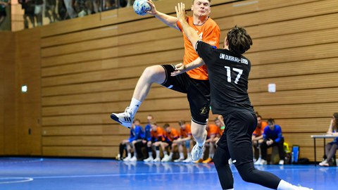 Foto: ein Sportler im orangenen Shirt springt in die Luft und holt mit dem rechten Arm, in der Hand ein Handball, zum Wurf aus; ein anderer Sportler versucht, den Wurf abzuwehren