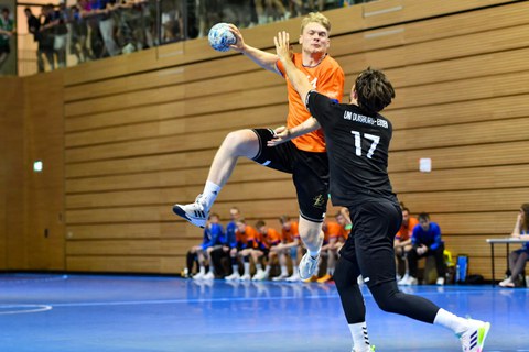 Foto: ein Sportler im orangenen Shirt springt in die Luft und holt mit dem rechten Arm, in der Hand ein Handball, zum Wurf aus; ein anderer Sportler versucht, den Wurf abzuwehren