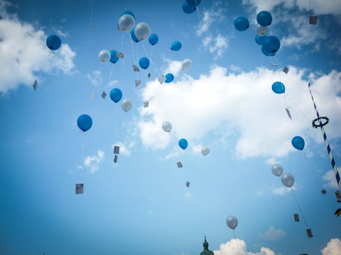 Das Foto zeigt zahlreiche graue und blaue Luftballons. Sie steigen in den blauen Himmel auf. An ihnen sind Postkarten befestigt. Am rechten Bildrand erkennt man zudem einen Maibaum.