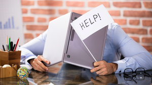 Foto eines Mannes am Schreibtisch. Er hält eine Fahne mit der Aufschrift "Help" und einen aufgeklappten Laptop in den Händen. Hinter diesem versteckt er seinen Kopf
