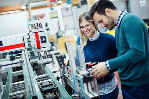 Das Foto zeigt zwei jüngere Menschen, die an einer hochtechnisierten Maschine stehen. Sie lächeln.