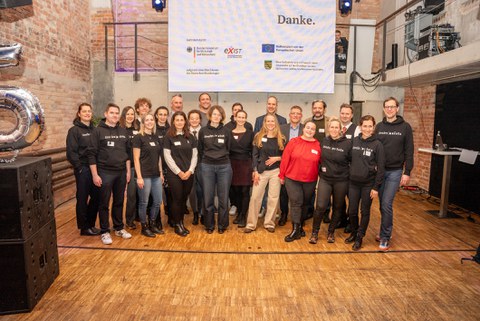 Gruppenfoto von circa 20 Personen in einem Veranstaltungssaal mit Industriestil, die Häfte trägt schwarze Shirts mit Aufdruck "dresden exists"