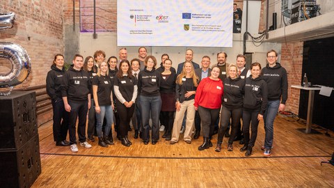 Gruppenfoto von circa 20 Personen in einem Veranstaltungssaal mit Industriestil, die Häfte trägt schwarze Shirts mit Aufdruck "dresden exists"