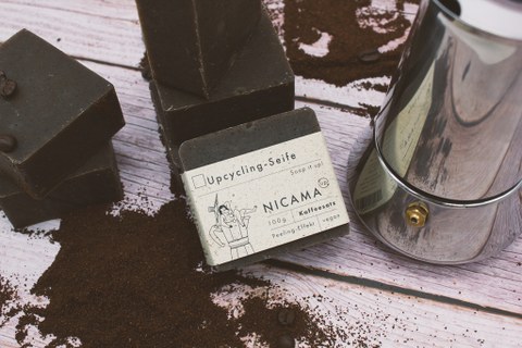 in der Mitte dunkle Seife beschriftet mit "Nicama", liegt auf Kaffeesatz, daneben ein Espressokocher