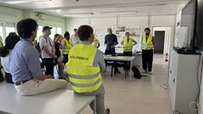 People standing or sitting in a semicircle listening to a presentation, some wearing yellow safety vests